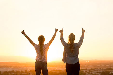 Two woman embracing a sunset and holding their thumbs up. 